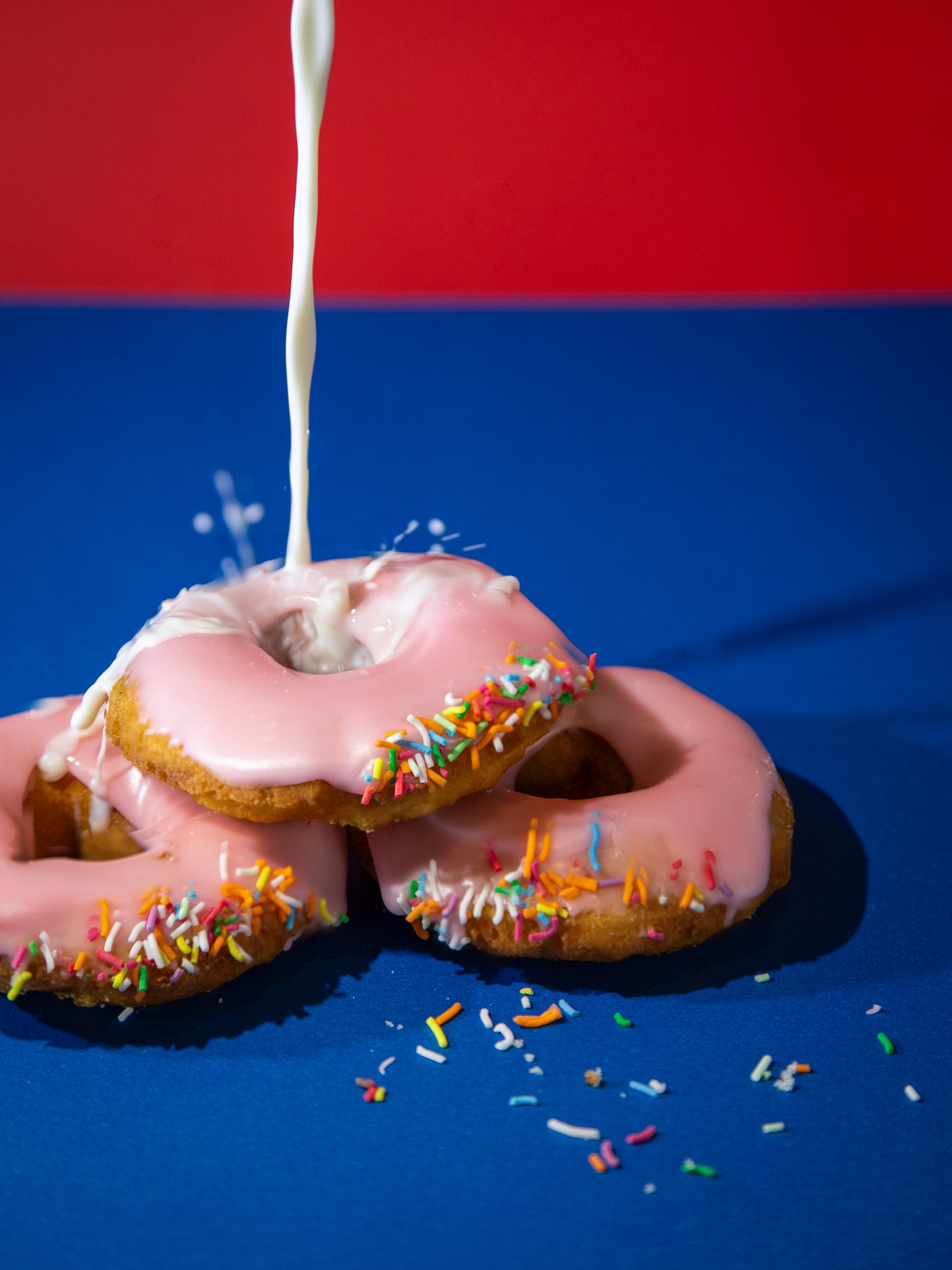 Photograph of fresh milk being poured over pink donuts against vibrant primary colour background. Upwey Village Bakery Donut Project photo shoot.