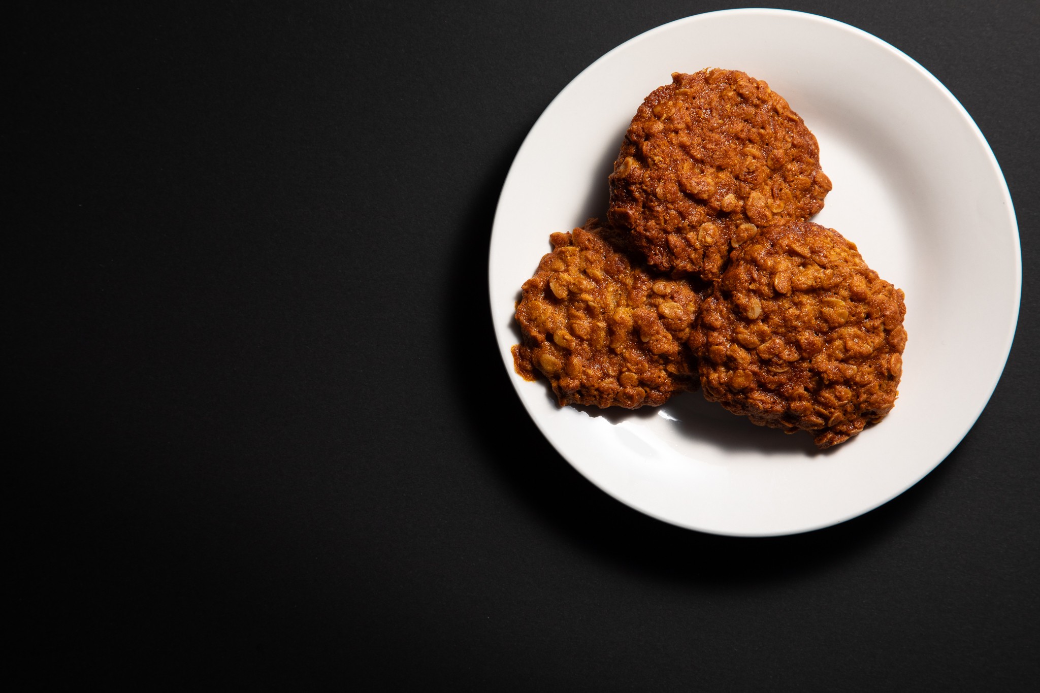 Flat lay food photograph of 3 Anzac biscuits on a white plate with black background. Negative space for editorial purposes